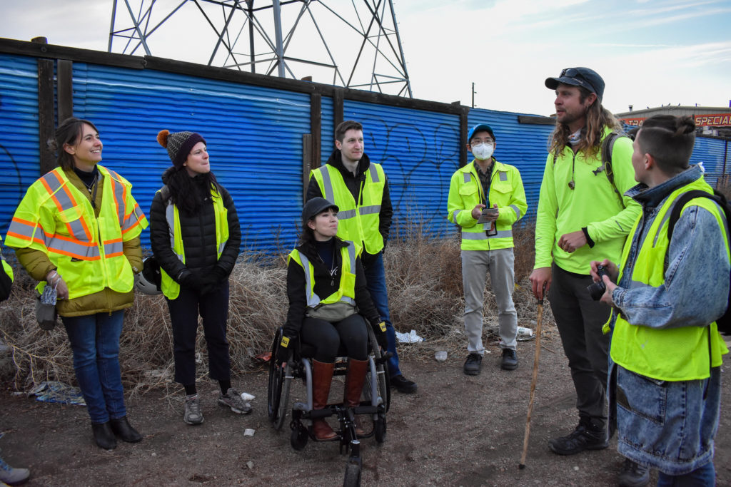 A group of people wearing yellow safety vests, standing and with one using a wheelchair, stand in a semi circle.