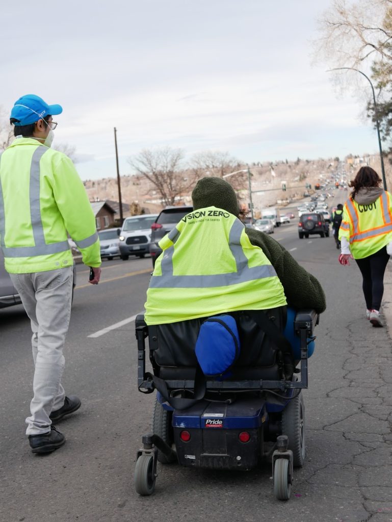 A person using a wheelchair with a yellow safety vest that says "Vision Zero" on it rolls in a driving lane of a road next to a person walking.