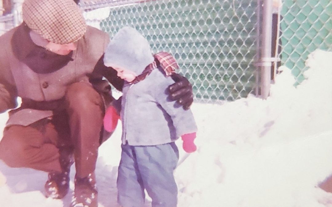 Jill as a young toddler standing on the snowy ground, and her father crouched next to her with his arm around her shoulder.