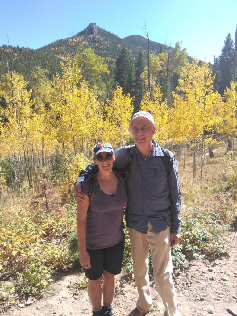 Jill posing with her father for a photo on a trail, in front of yellow aspens and a mountain.