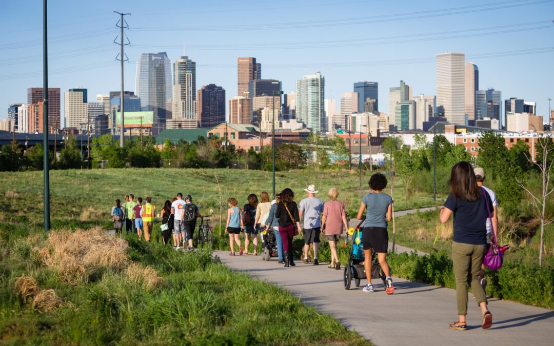 People walking on a paved trail in a grassy area with Denver's skyline in the background.