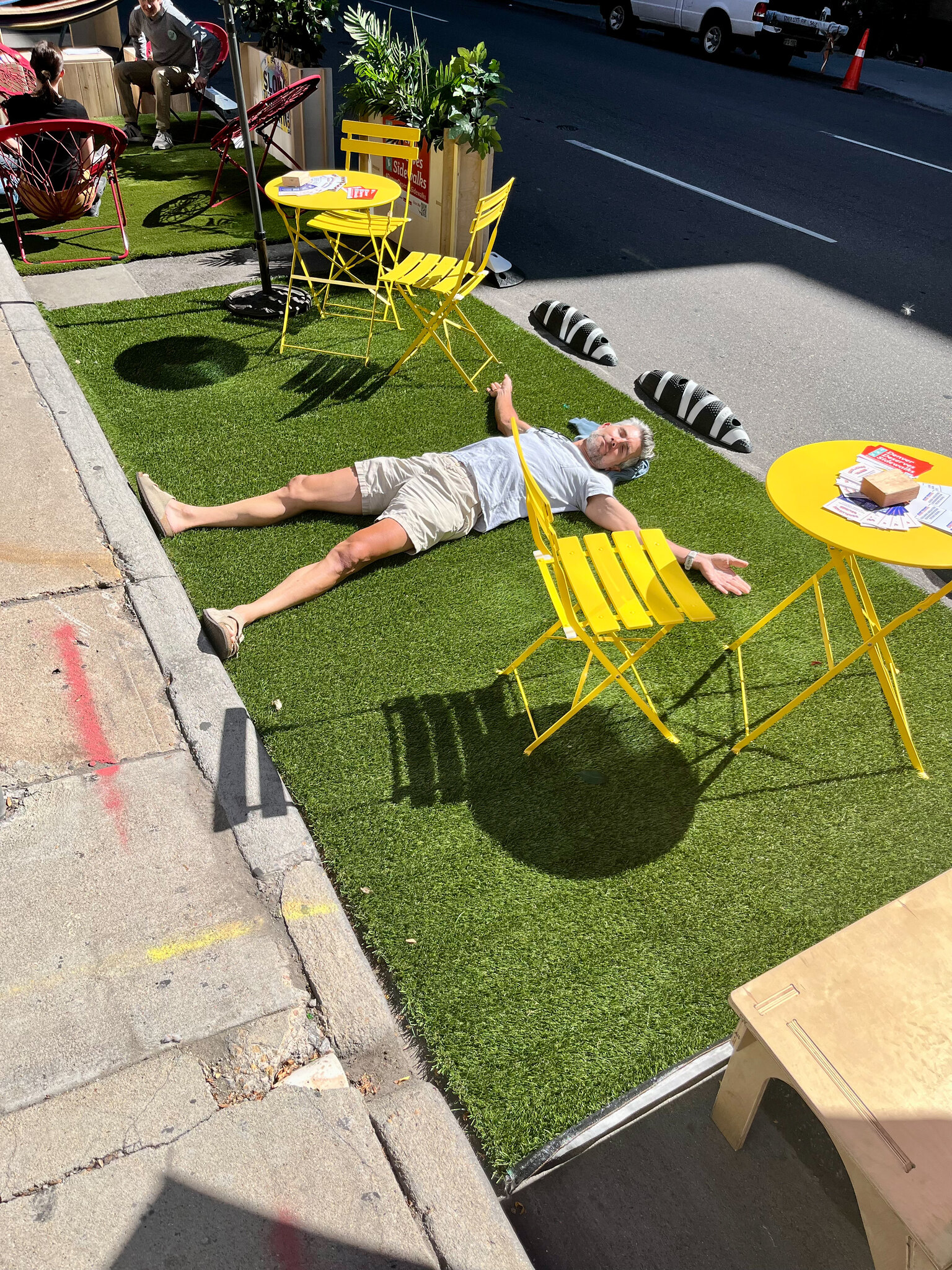 Pete Piccolo, a white man, lies flat on his back on a piece of turf in a street parking space that also has two small tables and chairs in it.