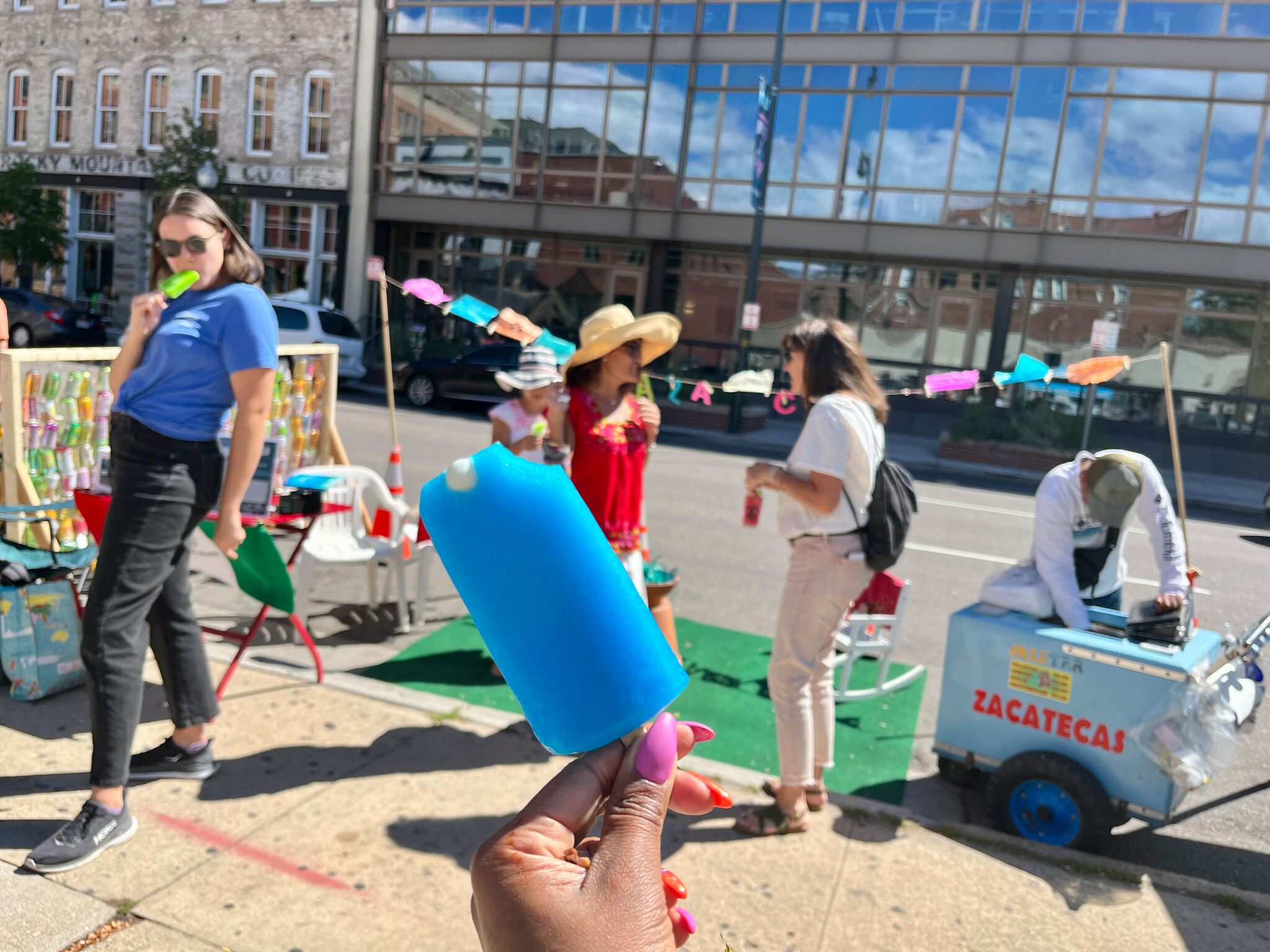 A hand holding a popsicle with a parklet in the background with a small popsicle cart and people enjoying.