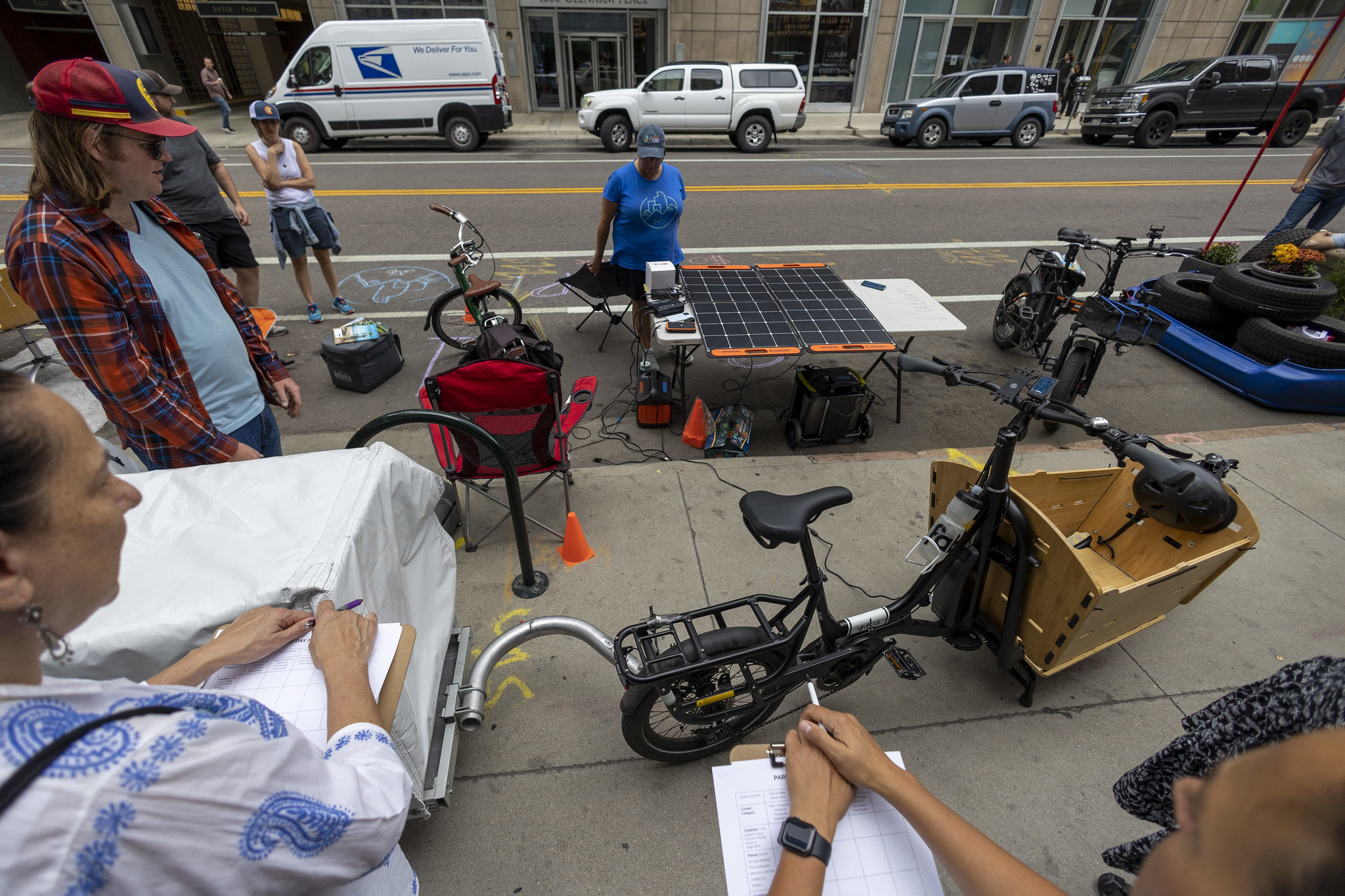 A solar-powered eBike charging station on a table in a street parking space. There are eBikes and people milling about.
