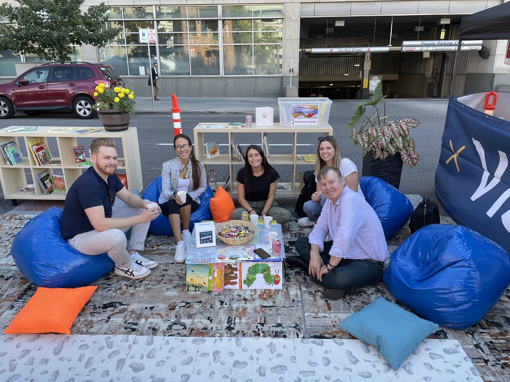 People sitting in a "living room" space with rugs on the floor and bookshelves lining a parking space on the street.
