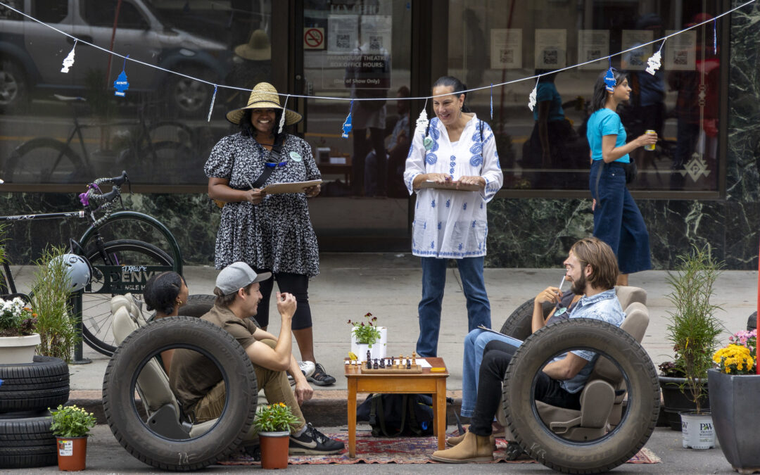 Four people sit on sofas made from the seats of a car, with armrests made from car tires, in a parking space on the street. Two people with clipboards stand facing them toward the camera.