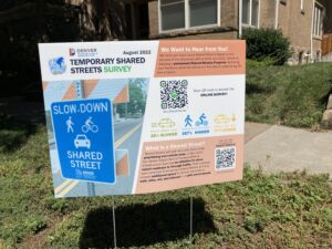 A yard sign reads, "Temporary Shared Streets Survey" with additional text, QR code and a photo of a "Slow Down Shared Street" sign.