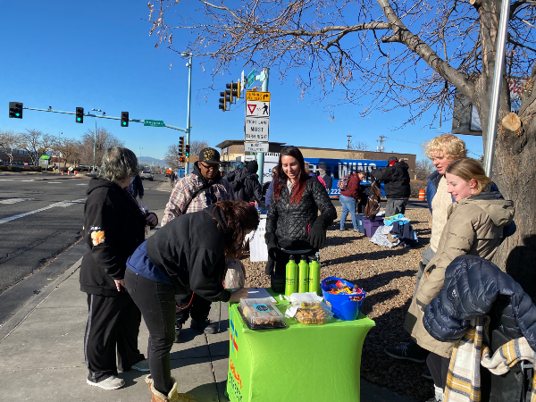 A few people gathering around a table next to a transit stop with treats and snacks on it.