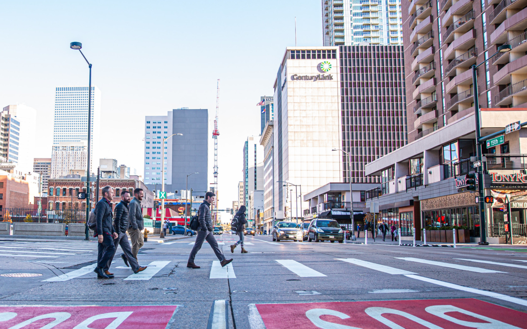 People crossing a city road with tall buildings surrounding them.