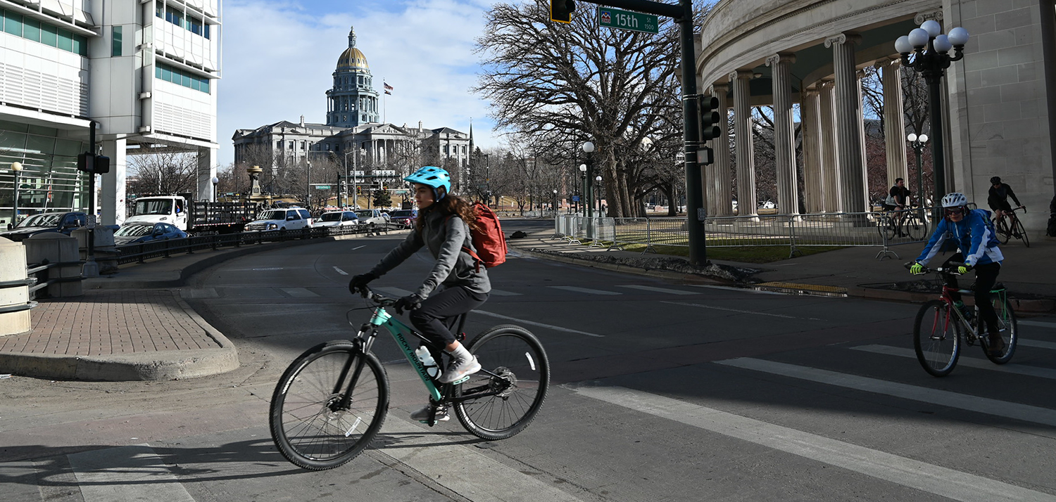 A bicyclist rides on a crosswalk underneath a traffic light, with the Denver capitol building in the background on a clear day.