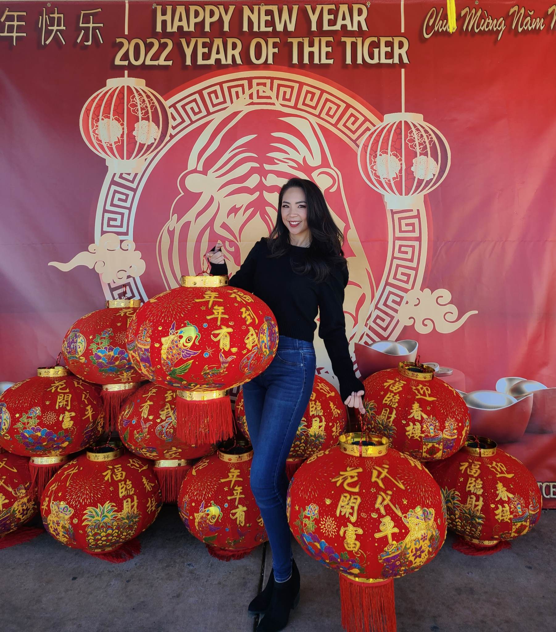 Mimi Luong stands in front of a banner wall titled, "Happy New Year: 2022 Year of the Tiger" and holds large, red cultural lanterns with gold writing in Chinese.