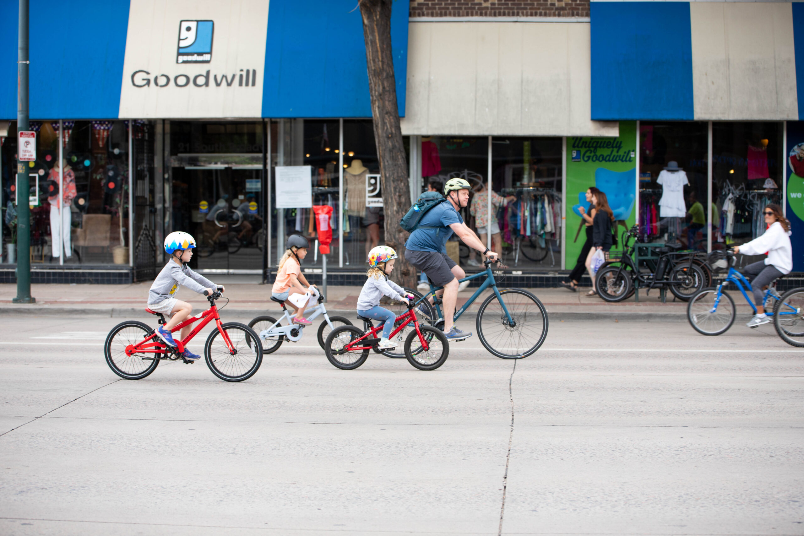 A dad and three kids bike up the South Broadway strip, passing in front of Goodwill, while the streets are closed to cars during ¡Viva! Streets 2024.
