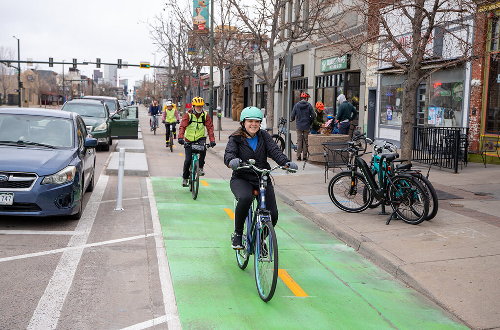 People smiling at the camera as they bike down the South Broadway bikeway, passing through a section where the lane is painted solid green.