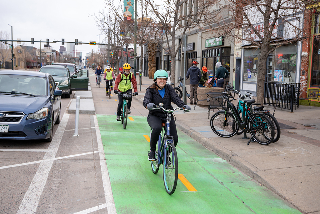 People smiling at the camera as they bike down the South Broadway bikeway, passing through a section where the lane is painted solid green.