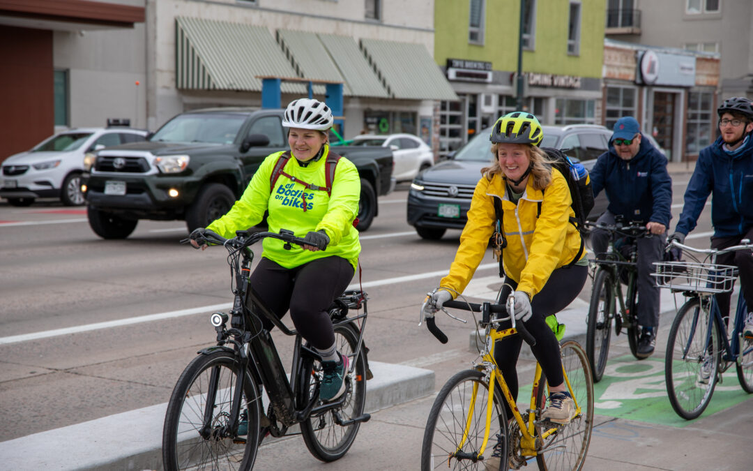 A group of bicyclists smile as they bike down the Broadway bikeway. A low concrete barrier separates them from vehicle traffic in the background.