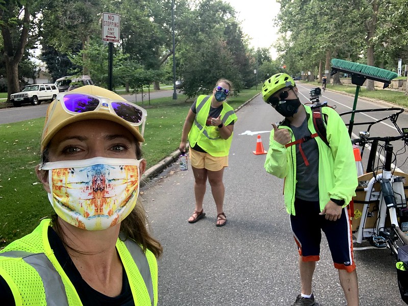 A woman takes a selfie on a street with two others giving thumbs up in the background. The three are wearing high-visibility gear and face masks. A bike lane, traffic cone, and cargo bike are in the background.