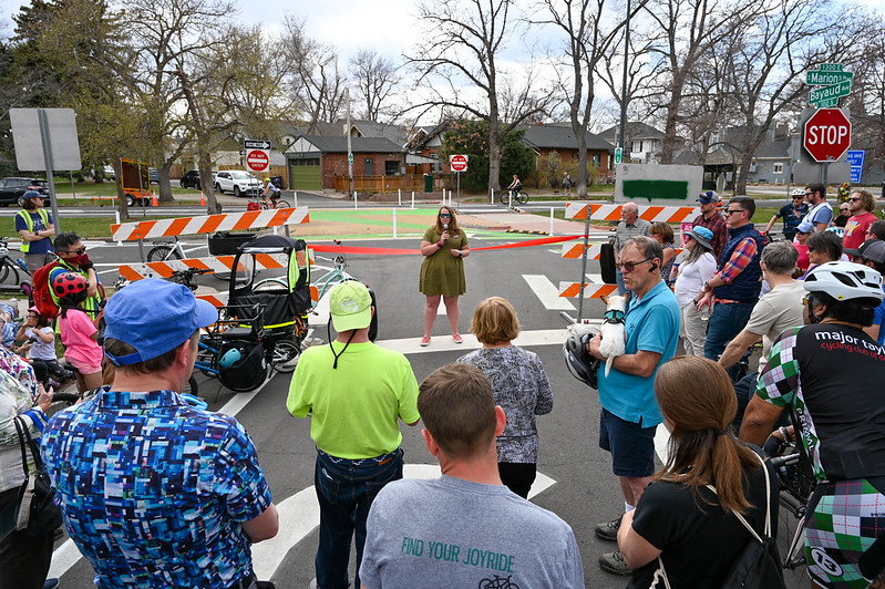 A woman with a microphone stands in front of a red ribbon marking the beginning of a bikeway. A crowd is gathered in the foreground.