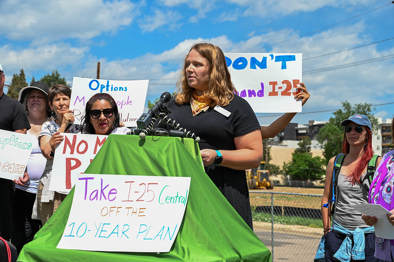 A woman speaks at a podium with an attached sign that reads "Take I-25 Central off the 10-year plan." A few others stand behind her, holding up additional handwritten signs.