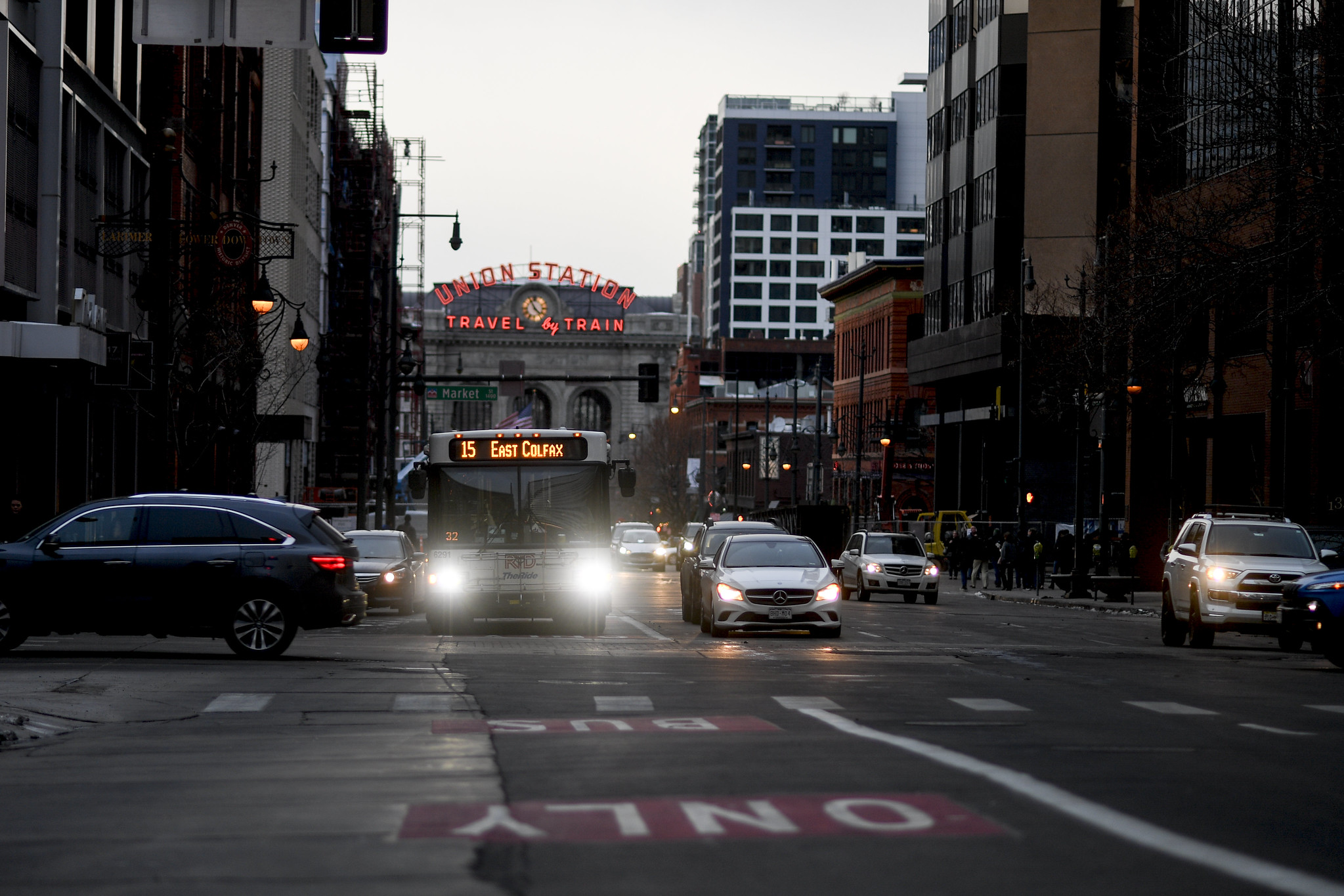 An RTD bus approaches the camera with bright headlights in a Bus Only Lane with Union Station behind it on an overcast day.