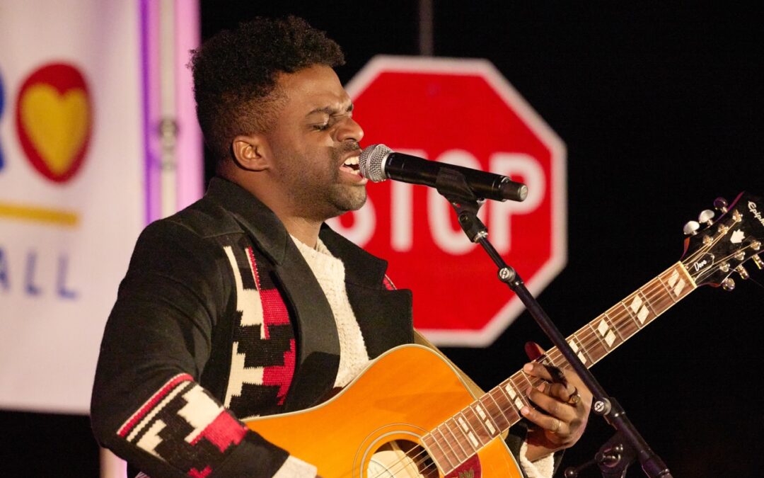 Close up of Abraham Alexander singing passionately into a microphone while playing an acoustic guitar.