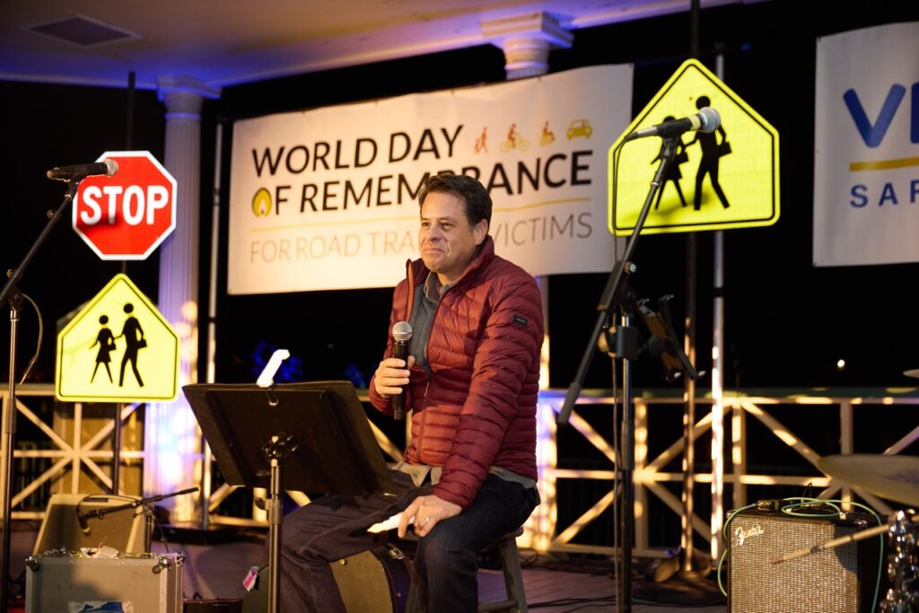 A man in a red jacket, Josh Stewart, speaks into a microphone he is holding as he sits on-stage. It's nighttime but the stage is well lit with reflective road signs and a banner that reads 