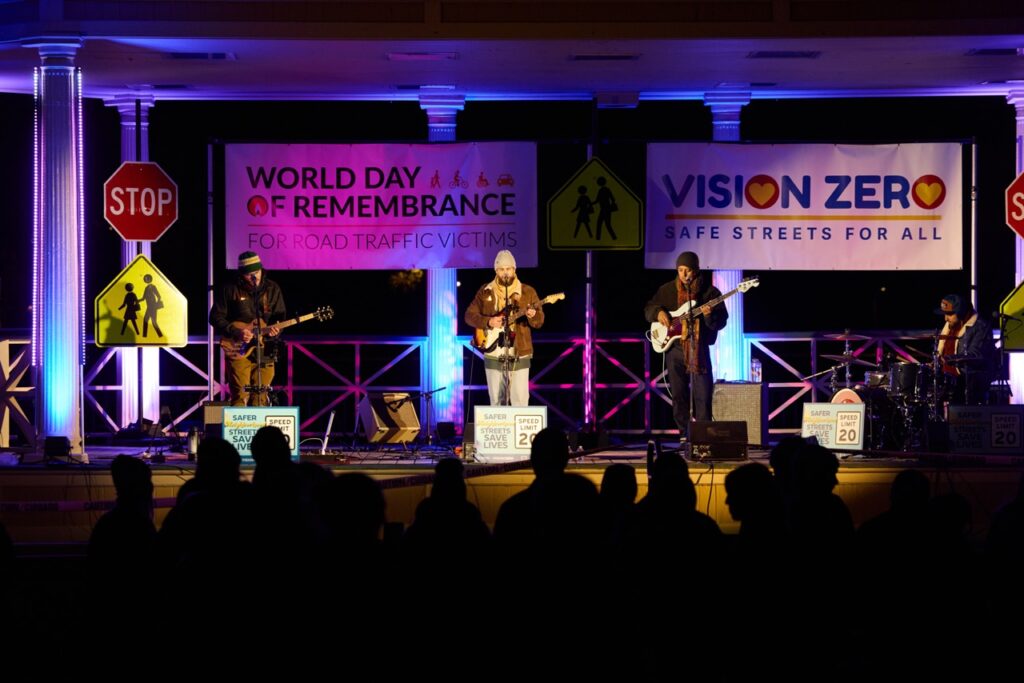 A silhouette of a crowd in the foreground faces a stage, where three men play guitars and sing, with a man to the right playing drums. Lights in hues of pink and blue light the stage and the road signs and banners behind it, which read 