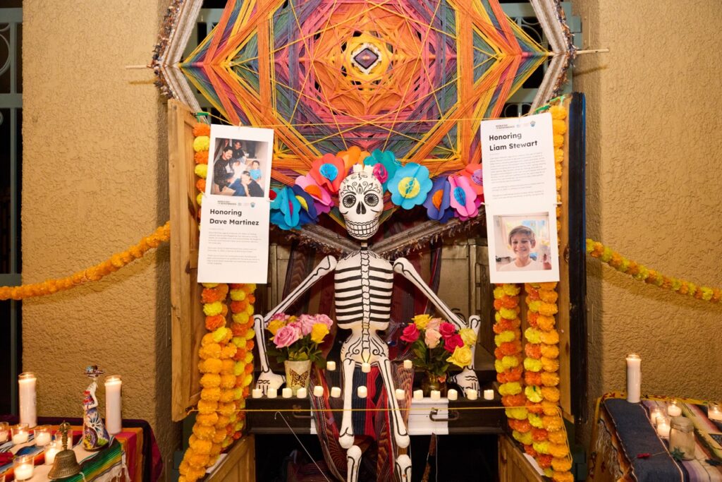 An ofrenda, or altar, contains a paper mache skeleton sitting in an open armoire that is strewn with strings of bright flowers and candles. A large yarn structure of colorful, geometric design is centered above the skeleton. Attached to the doors of the armoire are strings with papers clipped to them, featuring stories and photos of people's loved ones who have died in traffic crashes.