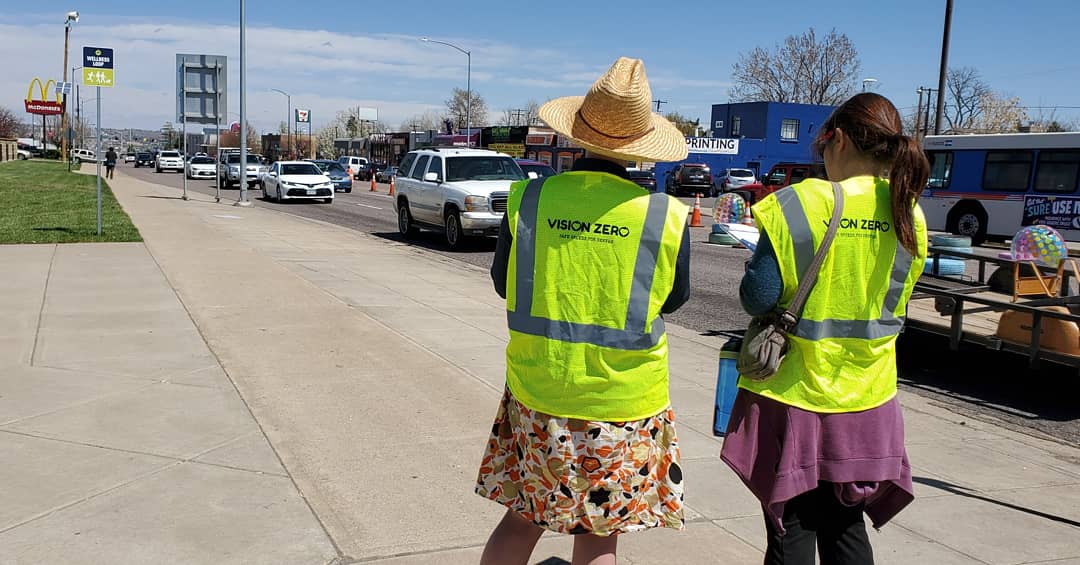 Two women wearing yellow safety vests with the Vision Zero logo stand on the side of Federal Boulevard