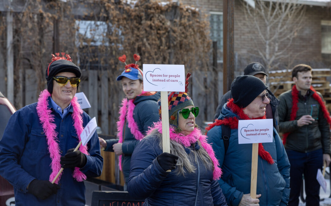 A group of people wearing red and pink boas and holding signs that say "spaces for people instead of cars"