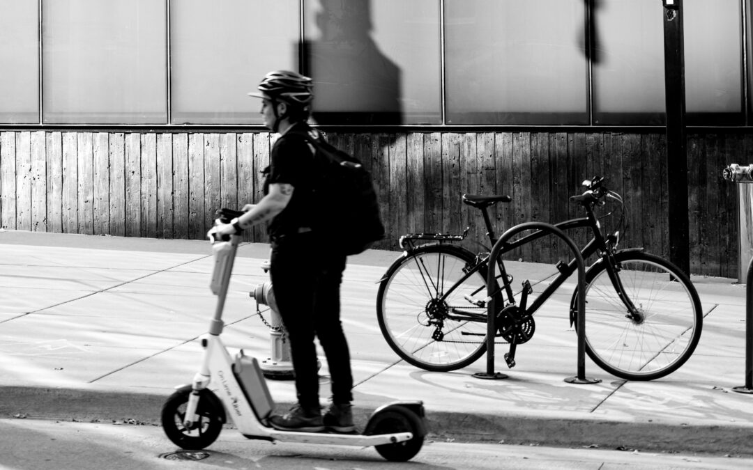 A person riding a stand-up e-scooter down the street past a bicycle locked to a bike rack