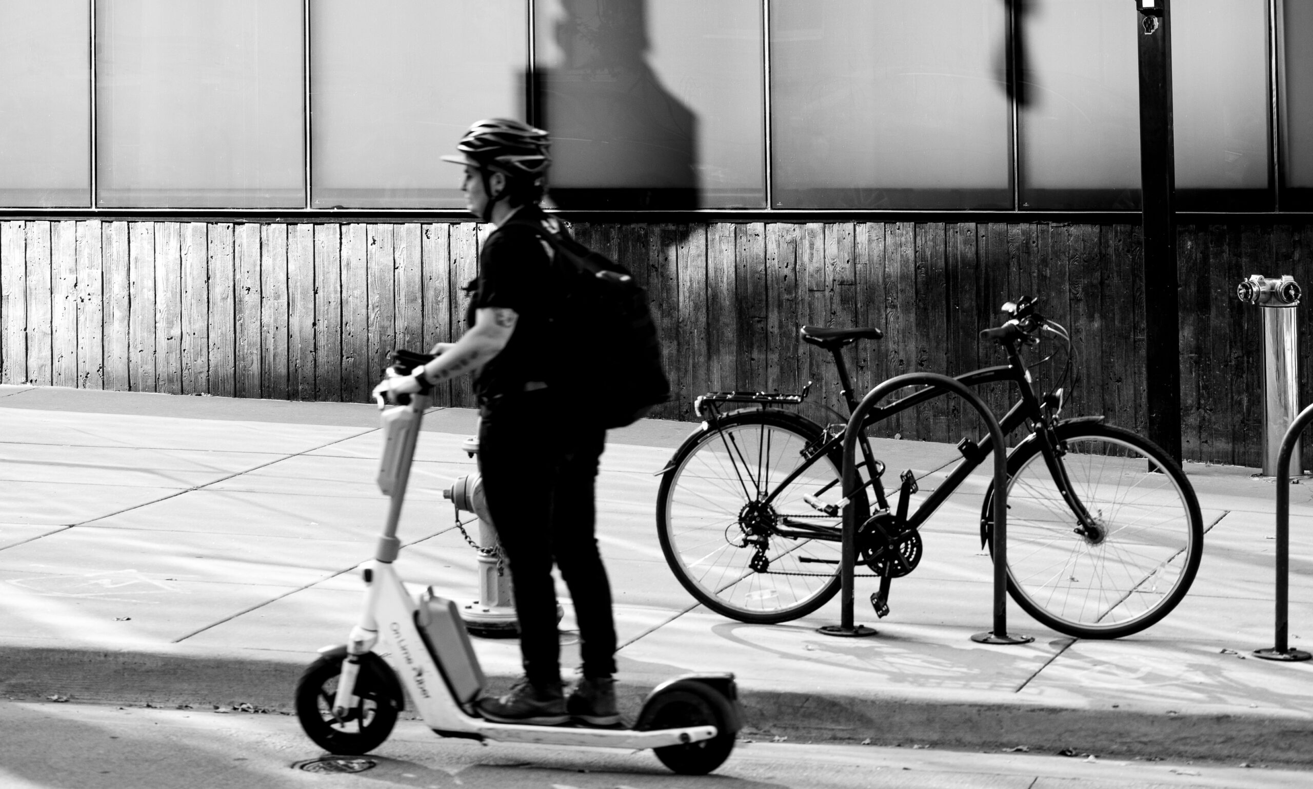 A person riding a stand-up e-scooter down the street past a bicycle locked to a bike rack
