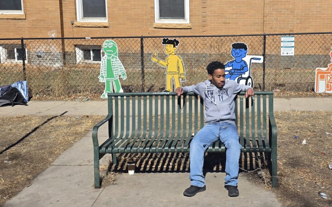 Artist AJ Kiel sits at the bus stop at MLK Jr. Blvd and York Street in front of his art installation, a series of colorful figures attached to a fence along the sidewalk