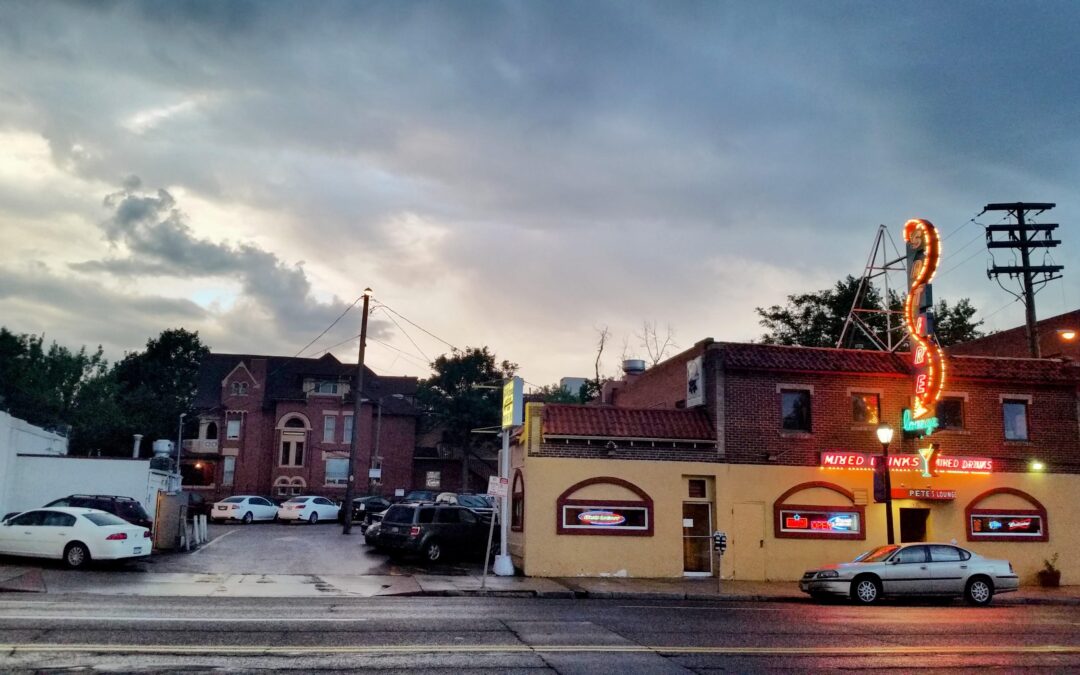 Photograph of East Colfax Avenue showing a single car parked on the street in front of Pete's Satire and an adjacent off-street parking lot