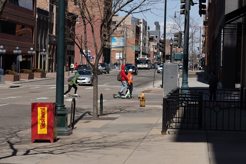 People walking, riding a scooter, and riding a skateboard on a busy street in downtown Denver